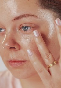 Nærbilde av en person som påfører klar gel hudpleieprodukt under øyet, med synlig blått �øye, lys hud og en gullring på fingeren.