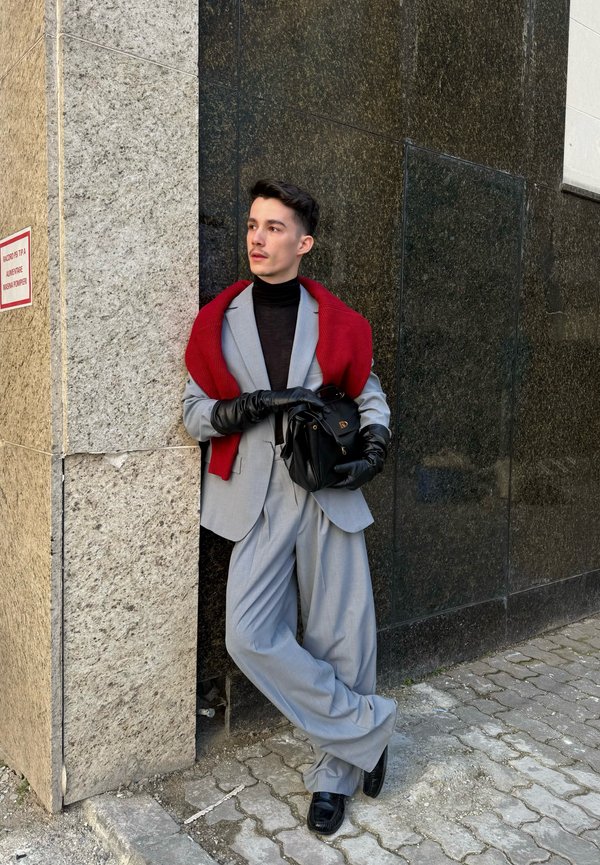 Giovane uomo in abito grigio chiaro e dolcevita nero appoggiato a un angolo di muro, indossa guanti neri e un maglione rosso drappeggiato sulle spalle.