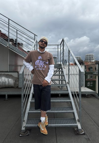 Camiseta gráfica marrón con estampado púrpura, llevada sobre una camisa blanca de botones, combinada con pantalones cortos de mezclilla oscura y botas beige con cordones amarillos, sobre escaleras de metal.