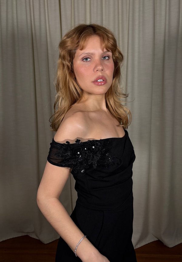 Jeune personne aux cheveux blonds ondulés portant une robe noire épaules dénudées avec des détails en dentelle florale, debout devant un rideau beige.