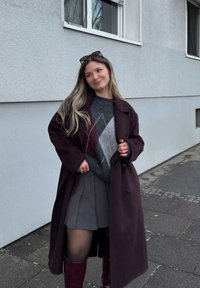 Jeune femme en long manteau sombre, pull à losanges, jupe plissée et bottes hautes se tient sur un trottoir près d'un bâtiment gris.