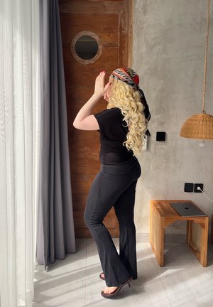 Mujer con cabello rubio largo y rizado, vestida de negro y con un pañuelo estampado en la cabeza, ajustándose cerca de una ventana con cortinas grises en una habitación moderna con paredes de madera y concreto.