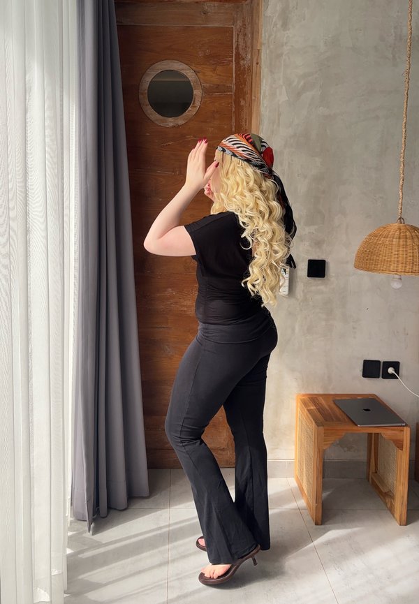 Žena s dlhými kučeravými blond vlasmi v čiernom oblečení a vzorovanej šatke na hlave si upravuje šatku pri okne so sivými závesmi v modernom priestore s drevenými a betónovými stenami.