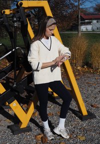 Cream v-neck sweater with a diamond pattern and navy accents, paired with dark leggings and white mid-calf socks, standing by gym equipment.