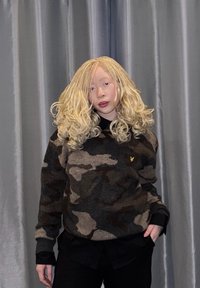 Le pull présente un motif camouflage en marron foncé, noir et beige, avec des poignets côtelés et un petit logo sur la poitrine. Chemise noire en dessous.