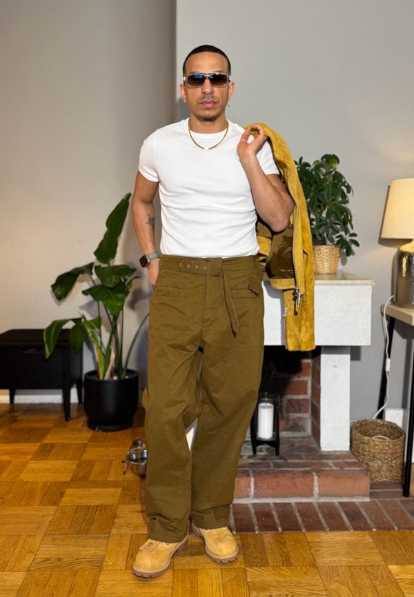 Uomo con occhiali da sole, maglietta bianca, pantaloni verde oliva e stivali color sabbia, che tiene una giacca gialla appoggiata su una spalla all'interno vicino a piante e un camino.