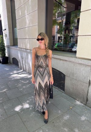 Un vestido maxi con un patrón en chevrón en tonos beige, negro y crema, con tirantes finos y una silueta fluida. Llevado con bailarinas.
