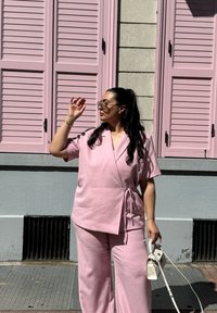 Lys pink indpakningsbluse med korte ærmer og matchende bredbensbukser. Modellen holder en lille hvid håndtaske, med en tekstureret baggrund af skodder.
