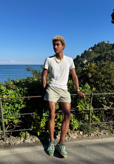 Camiseta blanca, pantalones cortos a rayas y zapatillas verdes. Collar con acentos naturales; de pie junto a una valla con vegetación y el mar de fondo.