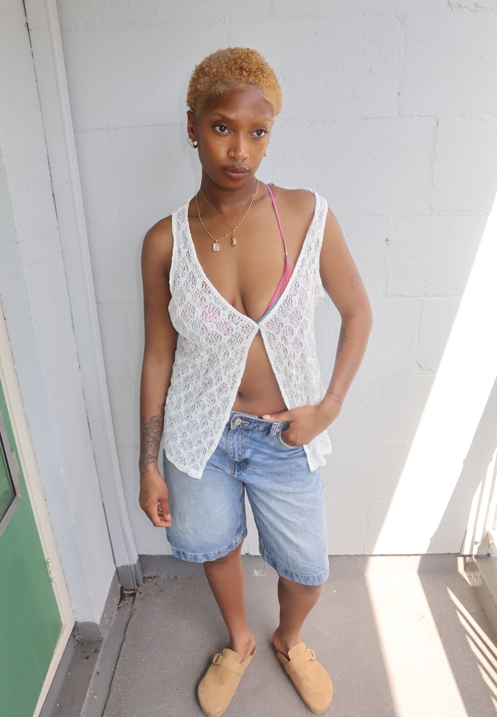 Mini top in pizzo bianco, pantaloni shorts di jeans, zoccoli beige. Pelle visibile sotto il top. Capelli corti e biondi, gioielli semplici, in piedi su una superficie di cemento.