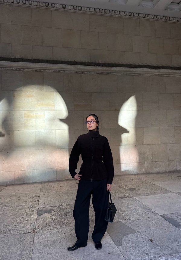 Una mujer vestida de negro está de pie con un bolso junto a un muro de piedra, enmarcada por dos brillantes manchas de luz solar.