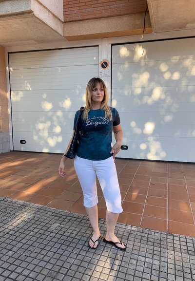 Mujer de pie en una acera con baldosas, vistiendo una camiseta oscura, pantalones capri blancos, chanclas negras y llevando un bolso negro de hombro.