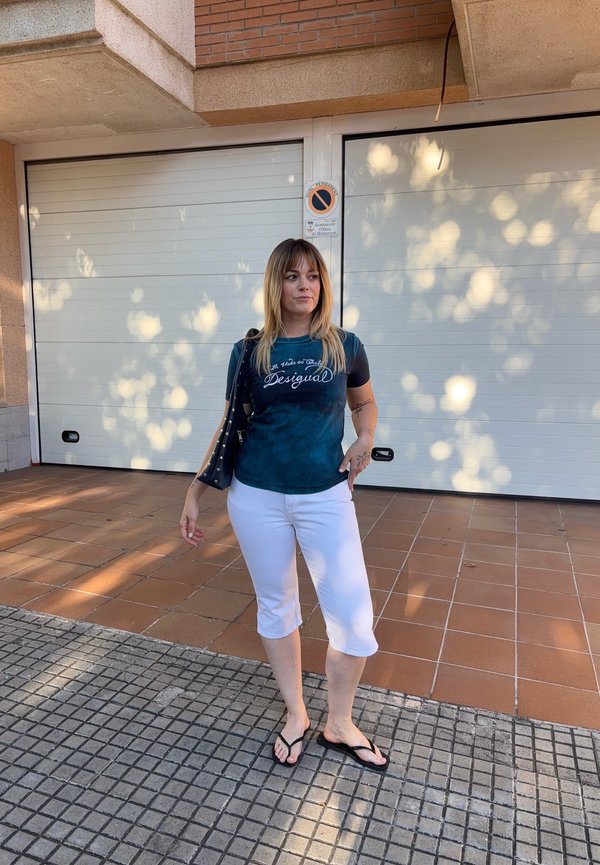Kvinde, der står på flisebelagt fortov iført en mørk t-shirt, hvide capri-bukser, sorte flip-flops og bærer en sort skuldertaske.