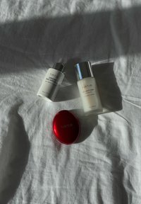 Produits de soin TIRTIR : une ampoule en céramique blanche au lait, une lotion tonique blanche avec des caps en argent, et un compact rouge brillant sur une surface blanche texturée.