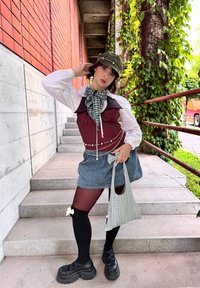 Rode polkadot top, witte blouse, spijkerrok. Zwarte knie-hoge kousen met strikjes, chunky zwarte schoenen, grijze hoed en geruite tas. Groene bladachtige achtergrond.