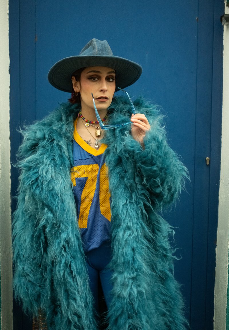 Personne portant un large chapeau bleu, un manteau duveteux bleu sarcelle, un maillot de sport bleu et jaune, tenant des lunettes de soleil bleues devant une porte bleue.