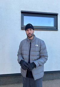 Casaco puffer cinza isolado com fecho de correr, luvas pretas e um gorro preto. Tecido suave e um logótipo na área do peito esquerdo.