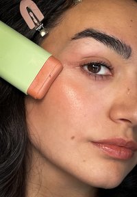 Mintgrüner Gesichtsroller mit einem pfirsichfarbenen Ende, das gegen die Haut gehalten wird. Das Gesicht des Models zeigt natürliches Make-up, dunkles Haar und definierte Augenbrauen.