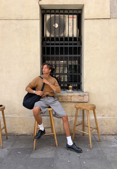 Suéter marrón de punto trenzado, bolso negro, pantalones cortos de cuadros, calcetines blancos, zapatos negros, sentado en un taburete de madera; el fondo presenta una ventana con rejas y un aire acondicionado.