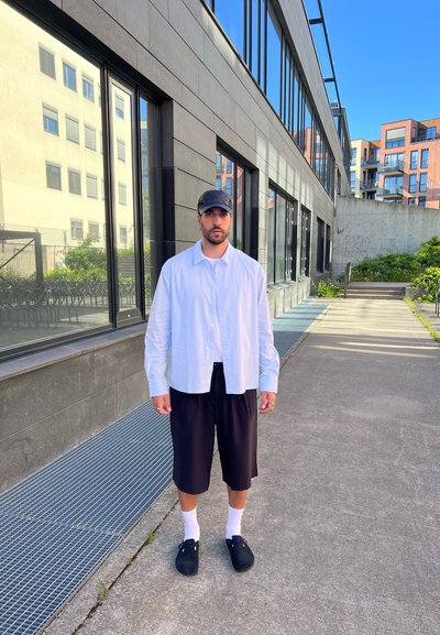 Camisa de botones blanca a rayas, pantalones cortos negros, calcetines blancos y zuecos negros. De pie cerca de un edificio moderno con grandes ventanas.