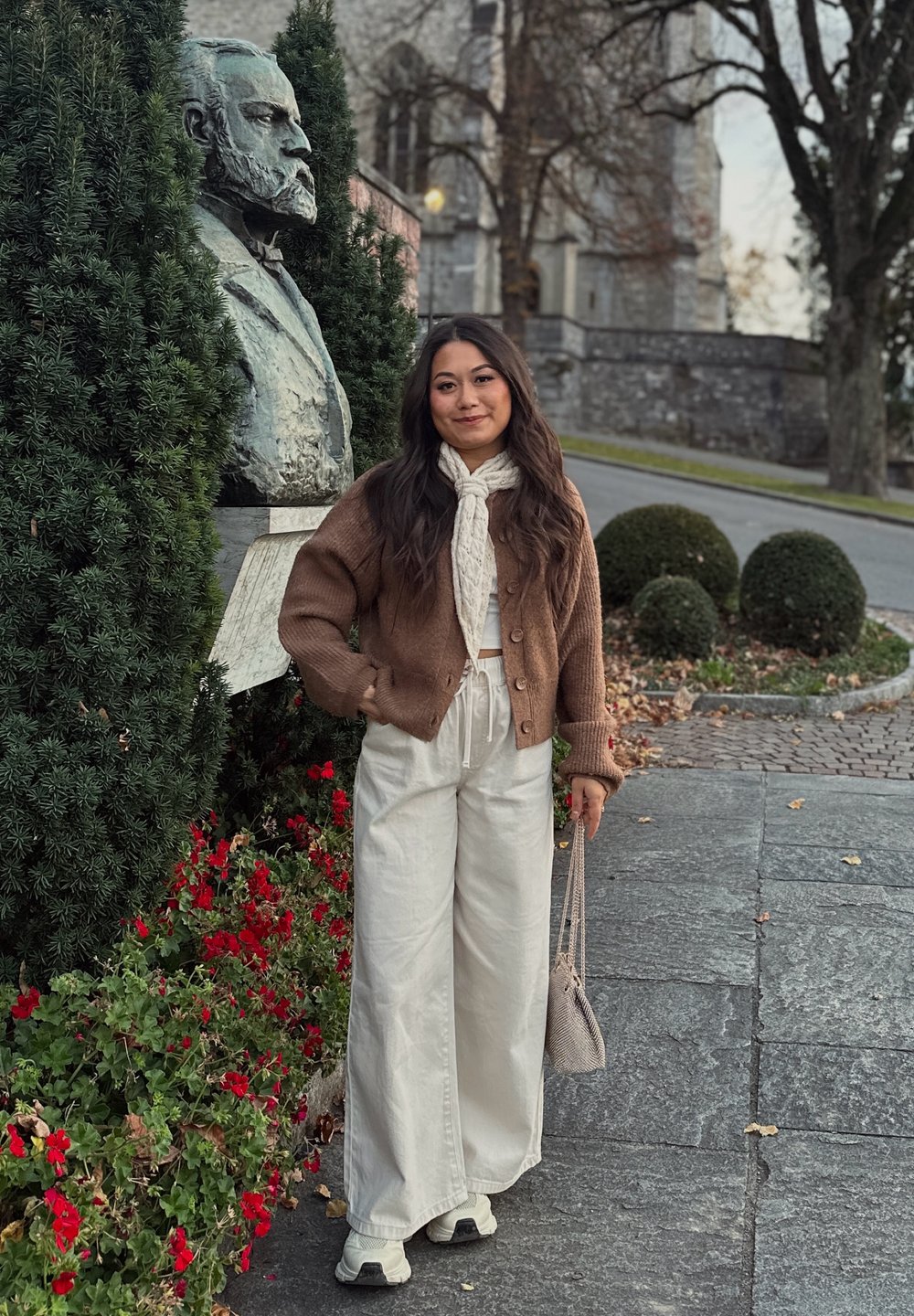 Hnědý pletený kardigan přes bílý top, v kombinaci se světle béžovými širokými kalhotami a teniskami, drží malou šedou kabelku, za níž je socha.