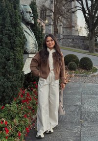 Hnědý pletený kardigan přes bílý top, v kombinaci se světle béžovými širokými kalhotami a teniskami, drží malou šedou kabelku, za níž je socha.