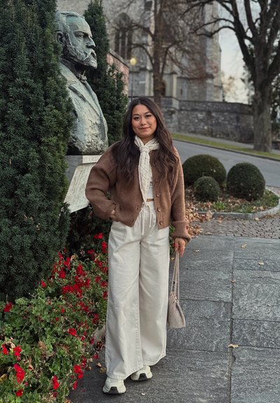 Cárdigan marrón de punto sobre una camiseta blanca, combinado con pantalones de pierna ancha de color beige claro y zapatillas deportivas, sosteniendo un bolso pequeño gris, con una estatua al fondo.