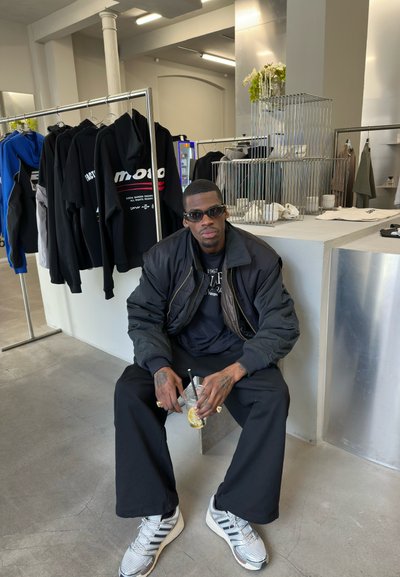 Hombre con chaqueta negra y gafas de sol sentado en el interior sosteniendo una bebida, rodeado de chaquetas colgadas y modernos accesorios de tienda.