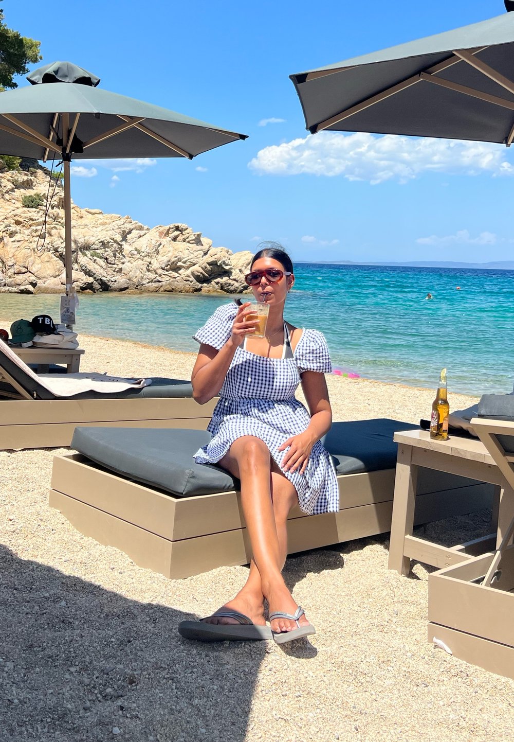 Karierter Kleid mit Puffärmeln, sitzt auf einem Liegestuhl und hält ein Getränk. Strandszene mit Sonnenschirmen, klarem blauen Wasser und felsiger Küste.