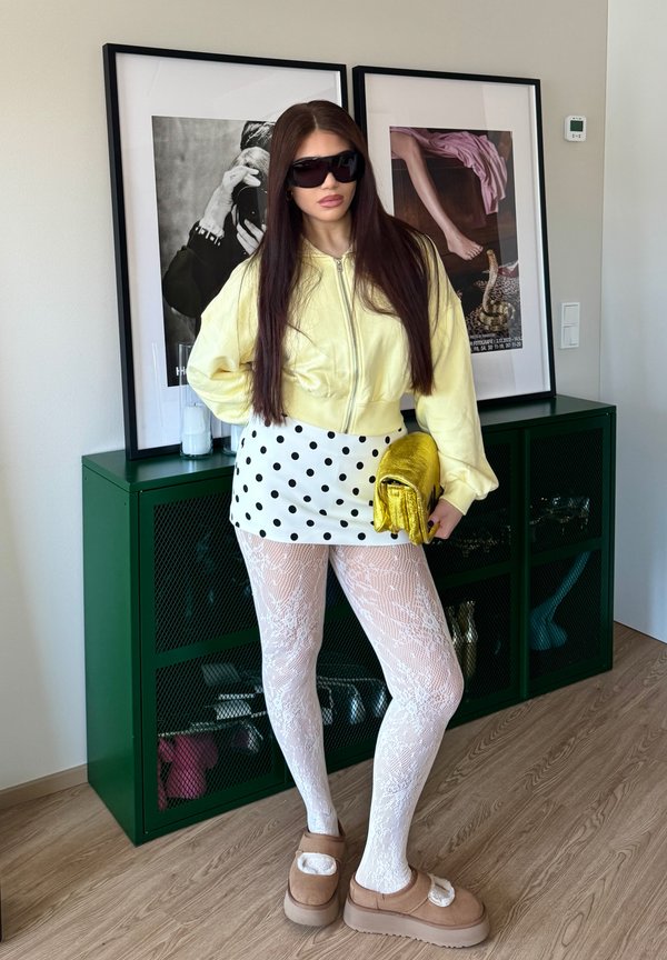 Kobieta w żółtej kurtce, spódnicy w groszki, białych koronkowych rajstopach, brązowych kapciach, trzymająca złotą kopertówkę, stoi w pomieszczeniu obok oprawionego dzieła sztuki.