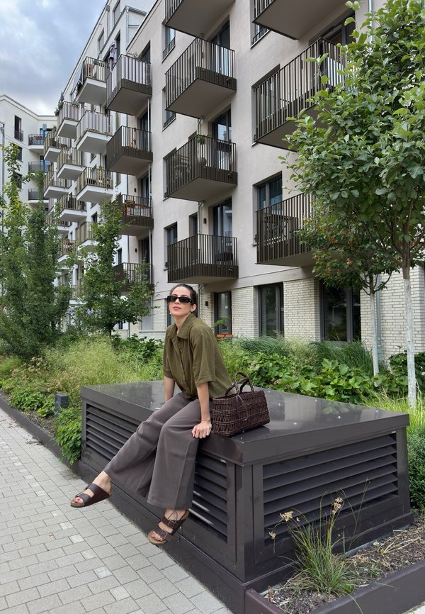 Femme portant des lunettes de soleil assise sur une structure métallique de ventilation devant des balcons d'appartements modernes avec de la verdure et des arbres à proximité.