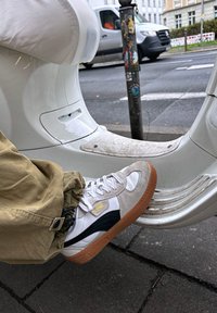 Balti ir pilki sportiniai bateliai su tekstūruotu paviršiumi, turi tamsiai mėlyną akcentą ir rudą guminę padą. Išdėstyti šalia baltos motorolerio.