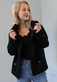 Black double-breasted coat with large buttons, paired with a black lace-trimmed top and patterned denim jeans featuring rhinestones.