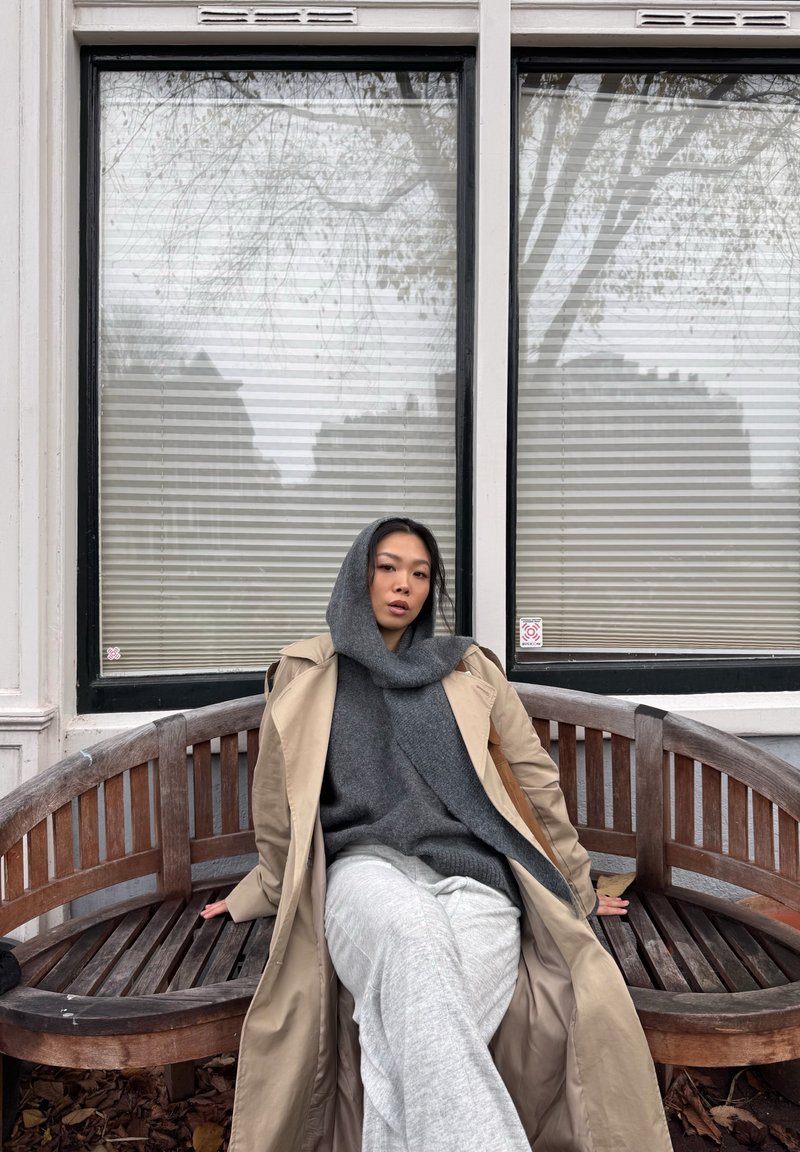 Una panca in legno con design a listelli sostiene una persona con un maglione grigio e pantaloni grigio chiaro, indossando un impermeabile beige, davanti a una finestra.