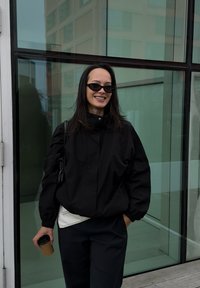 Jachetă neagră tip pulover, cu guler înalt, manșete strânse și o croială relaxată, asortată cu pantaloni bleumarin. Ținând o cană de cafea to-go maro.