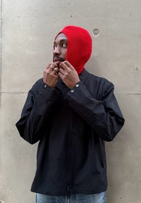 Fekete könnyű kabát patentos gombokkal, élénk piros kötött balaklávával páros�ítva. A modell a balaklávát igazítja, miközben balra néz.