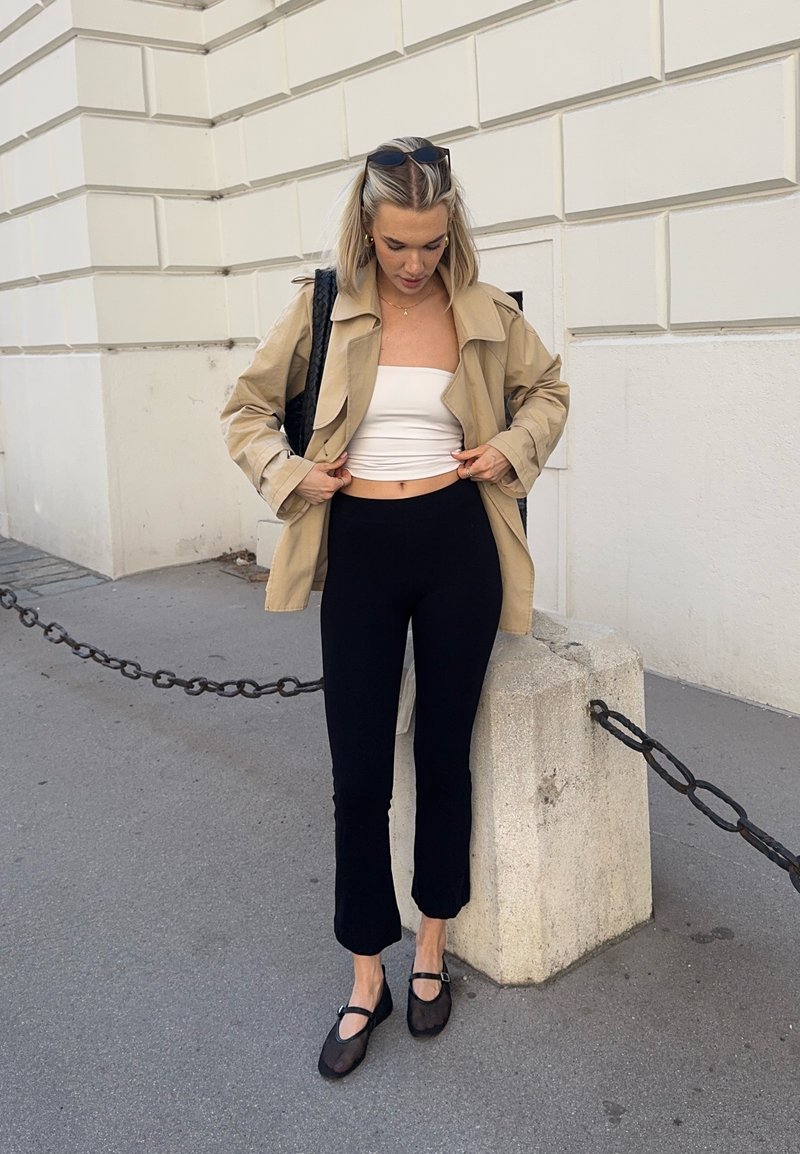 Jeune femme ajustant une veste beige, portant un crop top blanc, un pantalon noir, des chaussures noires et des lunettes de soleil sur la tête, debout sur le trottoir en ville.