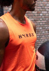 Narancssárga ujjatlan sportos trikó fekete "HYROX" logóval, sima anyagból készült és kerek nyakkivágással rendelkezik. Mellette fekete-fehér medicinlabda.