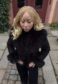 Čierna bunda s golierom a manžetami z umelého kožuchu, textúrovaná tkanina, dlhé rukávy, nosená s čiernymi nohavicami, na pozadí kamenného dlažobného kameňa.