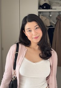 Helles pinkes Cardigan über einem weißen, gerippten Tanktop mit Spitzenrand. Schultertasche mit einem schwarzen Riemen und goldenen Beschlägen. Weiche, wellige Haare.