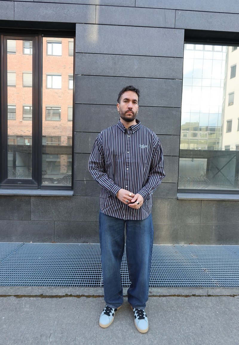 Homme debout devant un mur carrelé sombre, portant une chemise rayée à boutons, un jean ample et des baskets blanches avec des lacets noirs près des fenêtres d'un immeuble urbain.