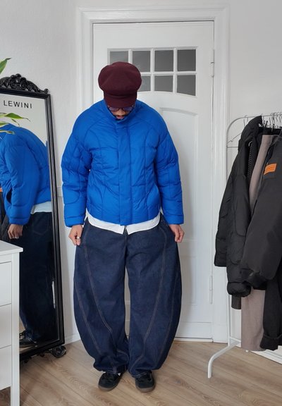 Chaqueta de plumas azul con cuello alto, combinada con pantalones de mezclilla oscuros de corte amplio. Debajo lleva una camisa blanca de botones. Zapatos negros.