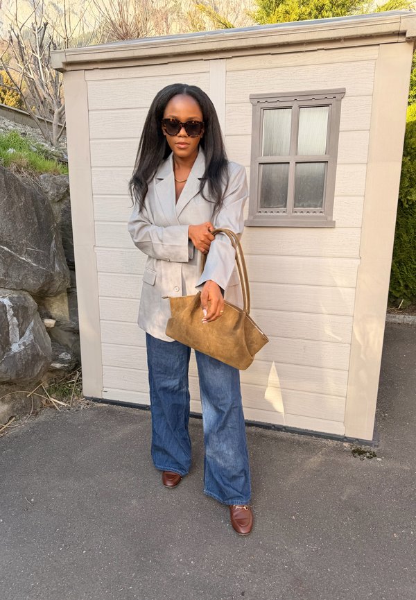 Donna con lunghi capelli neri, occhiali da sole, blazer grigio, jeans blu larghi, scarpe marroni e che tiene una grande borsa marrone davanti a una casetta chiara.