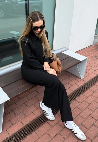 Mono negro de manga larga, de corte relajado, combinado con calcetines blancos y zapatillas grises. Un bolso marrón está colocado en el banco; el cabello largo peinado con clips.