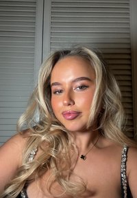 Femme blonde aux boucles lâches portant une robe avec bretelles étincelantes et un collier pendentif trèfle noir, devant une porte à persiennes.