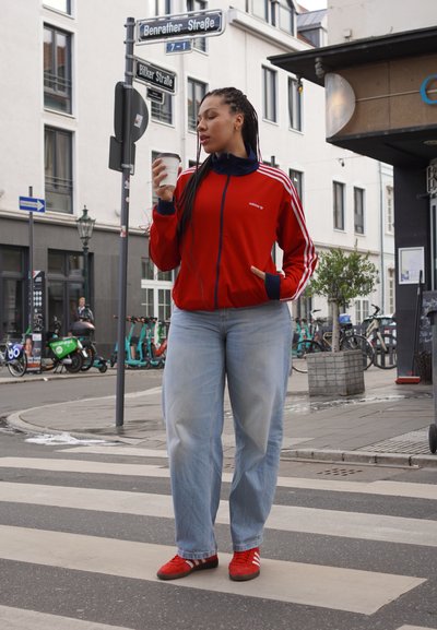 Chaqueta roja de Adidas con cremallera y franjas blancas, pantalones vaqueros holgados de color azul claro y zapatos rojos. Sosteniendo una taza de café blanca, de pie en un paso de peatones.