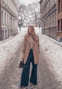 Femme en manteau beige et pantalon large noir debout dans une rue pavée bordée de neige, entre des bâtiments en brique et en pierre.