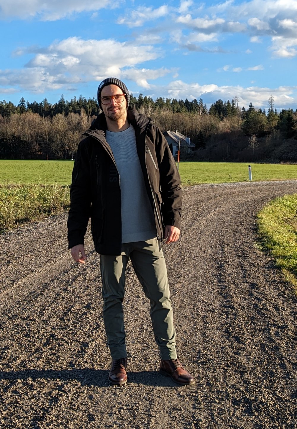 Zwarte jas, grijze shirt, groene broek, bruine laarzen. Staande op een grindpad omringd door groene velden en bomen onder een blauwe lucht.