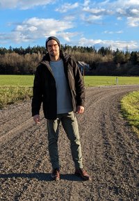 Zwarte jas, grijze shirt, groene broek, bruine laarzen. Staande op een grindpad omringd door groene velden en bomen onder een blauwe lucht.