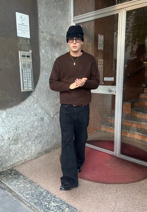 Suéter de punto marrón, vaqueros negros, gorro negro y gafas, de pie cerca de la entrada de un edificio con una pared de piedra y una alfombra circular en la entrada.